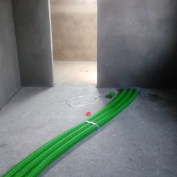 Control-AC Michał Machnik - Wnętrze w trakcie remontu z widocznymi, zielonymi rurami wentylacyjnymi, częściowo zwiniętymi w krąg, oraz trzema rurami wychodzącymi ze ściany, zabezpieczonymi taśmą. Surowe ściany i podłoga...