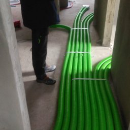 Control-AC Michał Machnik - Wiązka zielonych, karbowanych rur wentylacyjnych prowadzonych w surowym wnętrzu budynku, zabezpieczonych szarymi opaskami zaciskowymi, widoczna osoba w ciemnym ubraniu.