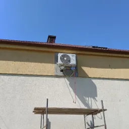 Zewnętrzna jednostka klimatyzacji zamontowana na elewacji budynku, widoczne podłączenia przewodów i rusztowanie.