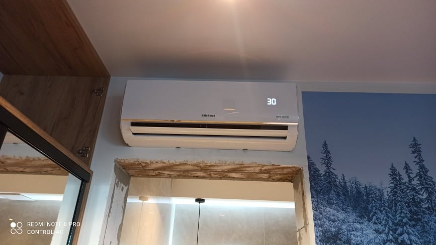 Biała jednostka klimatyzacji Samsung Digital Inverter zamontowana na ścianie w pomieszczeniu z tapetą w motywie zimowego lasu, ustawiona na temperaturę 30 stopni Celsjusza.