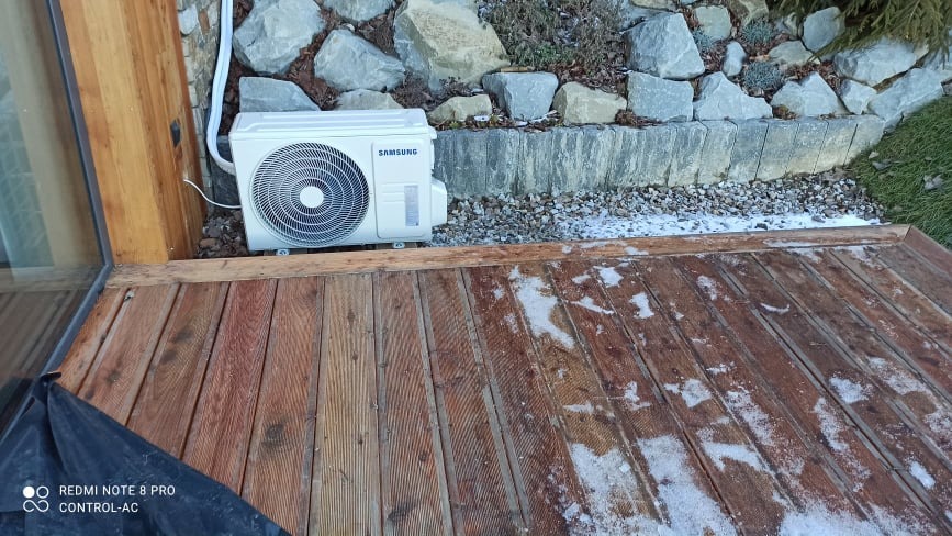 Zewnętrzna jednostka klimatyzacji Samsung zamontowana na drewnianym tarasie, obok ściany budynku, z kamiennym obrzeżem w tle, lekko przyprószona śniegiem.