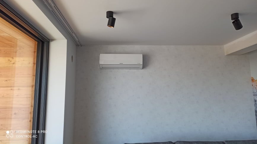 Biała jednostka klimatyzacji zamontowana na jasnej ścianie w nowoczesnym wnętrzu, widoczne czarne reflektory sufitowe i fragment drewnianej ściany.