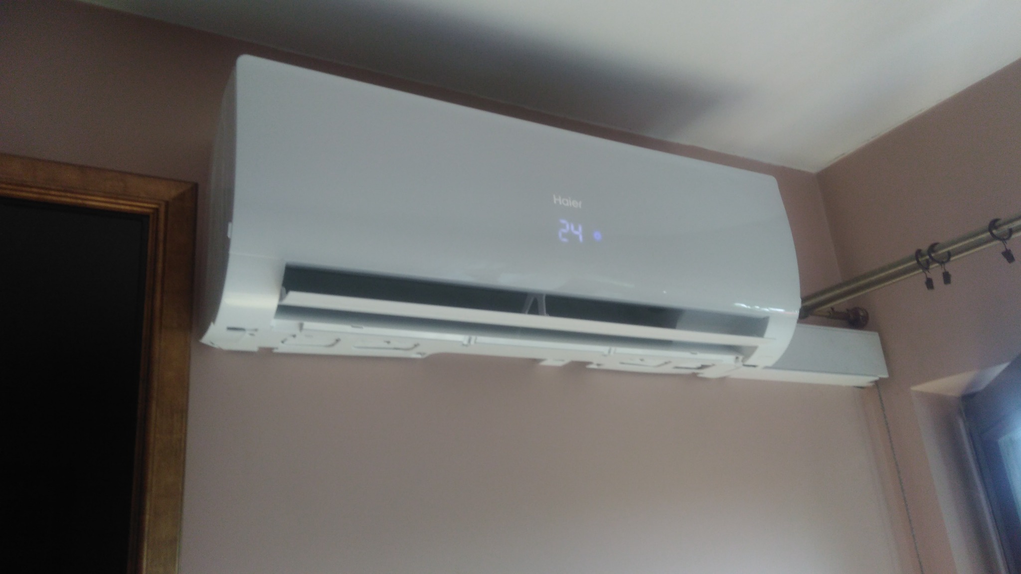 Biały klimatyzator Haier zamontowany na ścianie o odcieniu beżowym, widoczny wyświetlacz z temperaturą 24 stopnie, obok złota rama obrazu i karnisz z zasłonami.