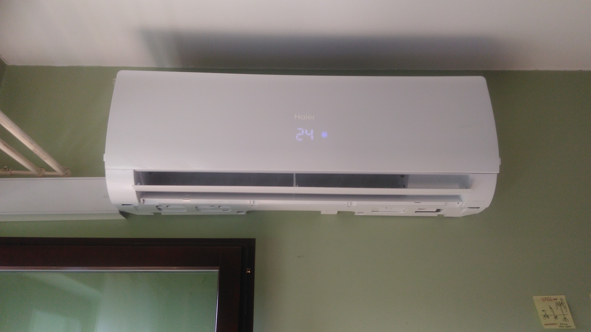 Biała jednostka wewnętrzna klimatyzatora Haier zamontowana na zielonej ścianie, widoczny wyświetlacz z temperaturą 24 stopnie, obok suszarka na pranie i fragment okna.