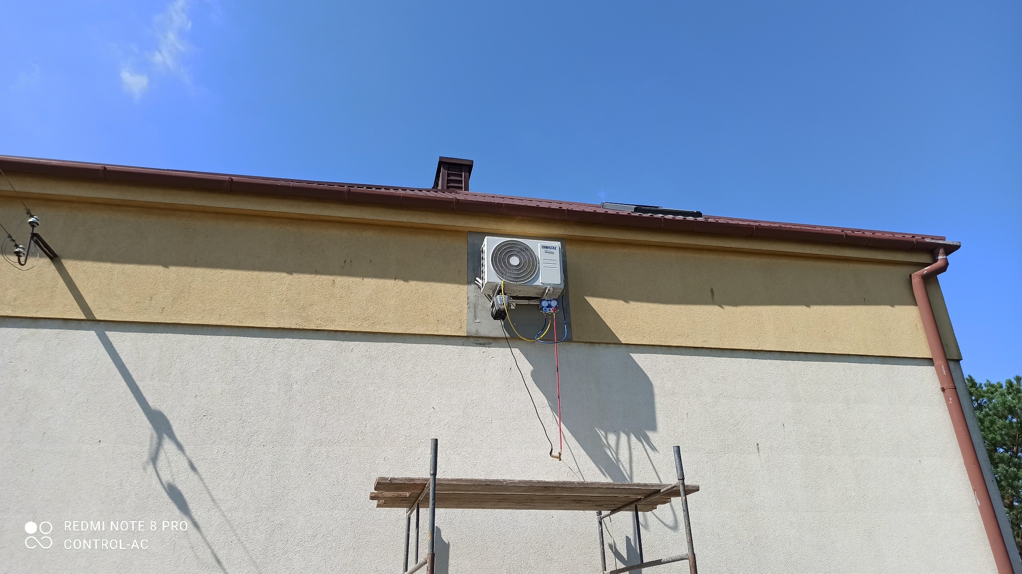 Zewnętrzna jednostka klimatyzacji zamontowana na elewacji budynku, widoczne podłączenia przewodów i rusztowanie.