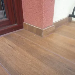 Ułożona podłoga z płytek imitujących drewno na balkonie, z cokolem dopasowanym do koloru podłogi, obok filaru elewacyjnego w kolorze terakoty i ramy okiennej w kolorze mahoniu.
