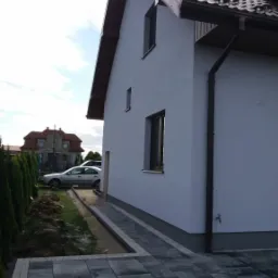 Świeżo otynkowany dom jednorodzinny z grafitową dachówką i rynnami, widok z boku na elewację i chodnik z kostki brukowej, w tle drugi dom i zaparkowany samochód.