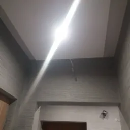 Wykończone wnętrze: Szpachlowane ściany z poziomą teksturą w odcieniach szarości, biały sufit z obniżonym fragmentem i wystającym kablem, widoczny fragment drewnianych drzwi.