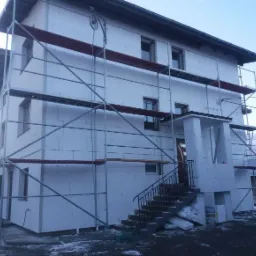 Dwupiętrowy dom w trakcie ocieplania, pokryty białymi płytami styropianowymi, otoczony metalowym rusztowaniem, z widocznymi schodami wejściowymi i fragmentem podwórka z resztkami śniegu.