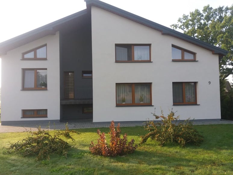 Dwukondygnacyjny dom z białą elewacją i szarym wnęką, drewniane okna, numer domu '9' widoczny na ścianie, zadbany trawnik z krzewami.