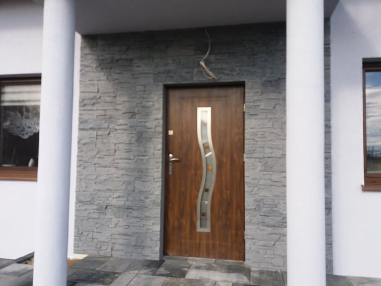 Wejście do domu w Brzeźniu z drewnianymi drzwiami z dekoracyjnym przeszkleniem, obłożone szarą kamienną elewacją, widoczne białe kolumny.