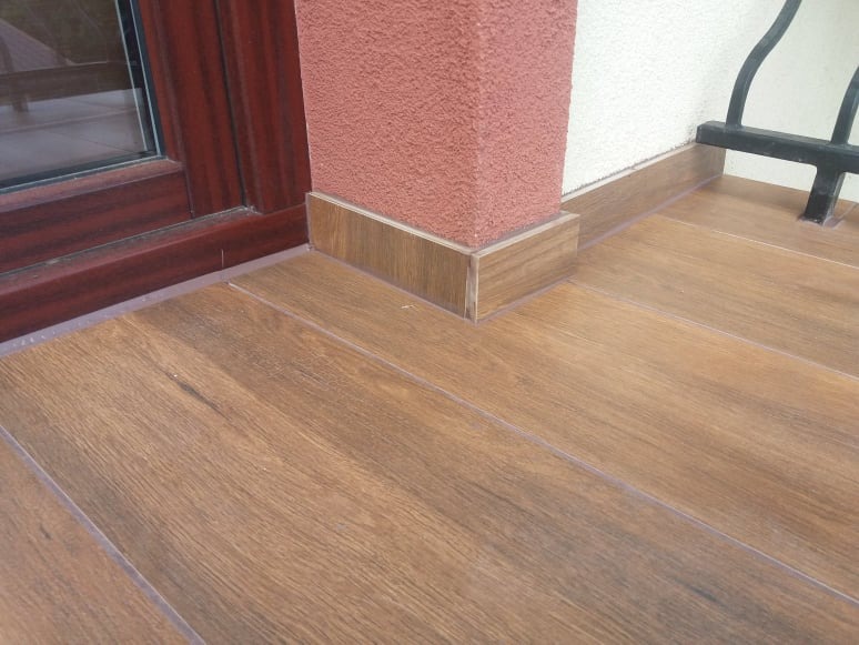 Ułożona podłoga z płytek imitujących drewno na balkonie, z cokolem dopasowanym do koloru podłogi, obok filaru elewacyjnego w kolorze terakoty i ramy okiennej w kolorze mahoniu.