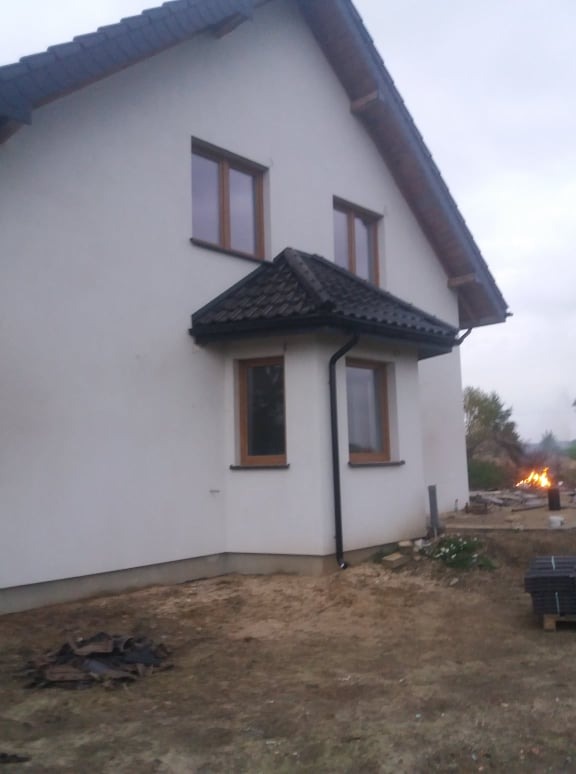 Biały dom jednorodzinny z brązowymi oknami i ciemną dachówką, widoczne prace wykończeniowe na zewnątrz, w tle ognisko i materiały budowlane.