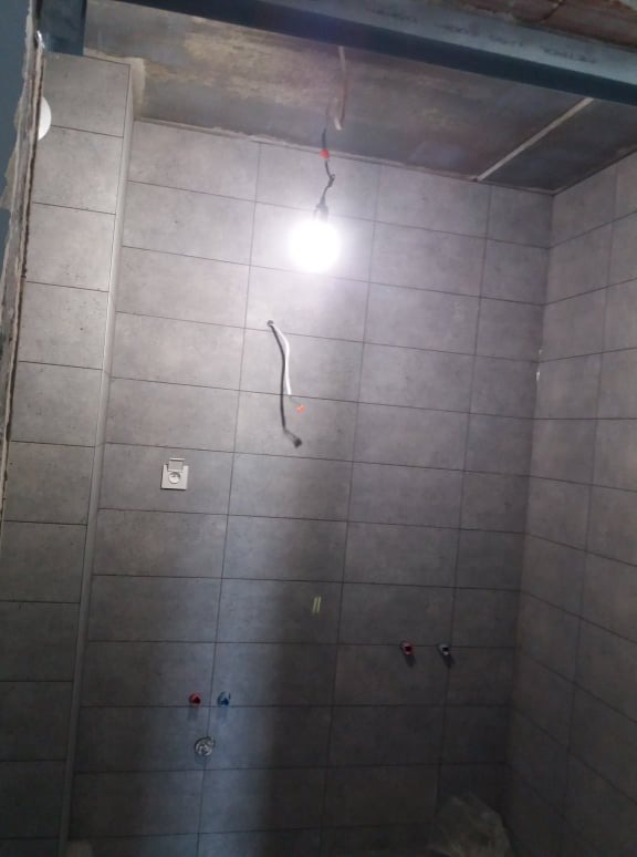Ściana łazienki w trakcie układania szarych płytek, widoczne wystające rury instalacji wodnej i elektrycznej, surowy betonowy sufit z prowizorycznym oświetleniem.
