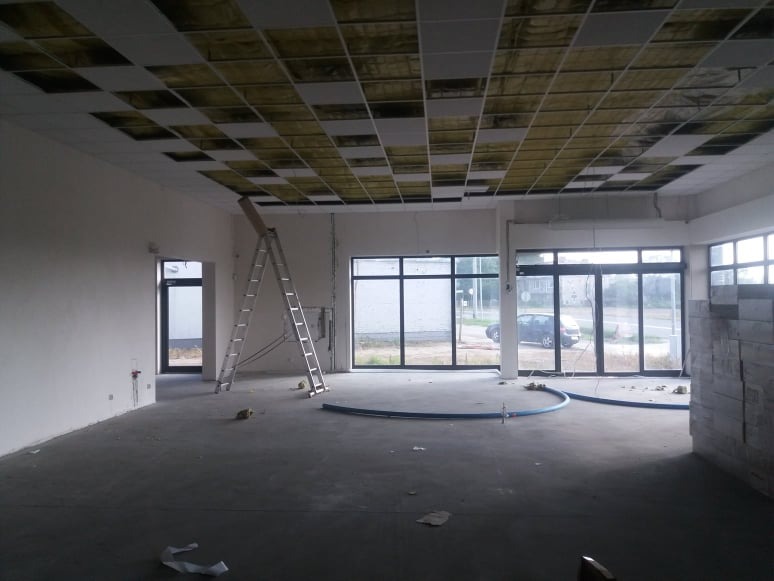 Wnętrze w trakcie wykończenia: widoczny sufit podwieszany z częściowo odsłoniętą izolacją, drabina oparta o ścianę, duże okna i niebieski przewód na szarej podłodze.