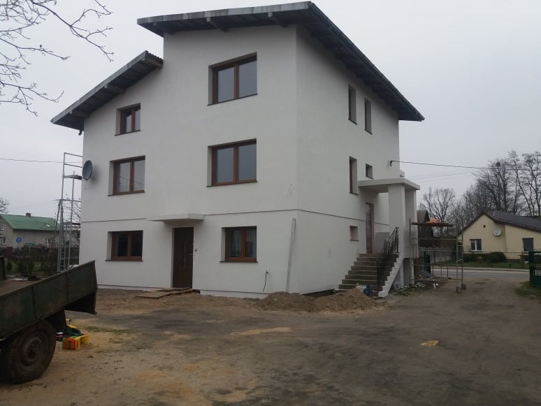 Dwupiętrowy dom z nową, białą elewacją i brązowymi oknami, widoczne rusztowania i teren wokół domu w trakcie prac wykończeniowych.