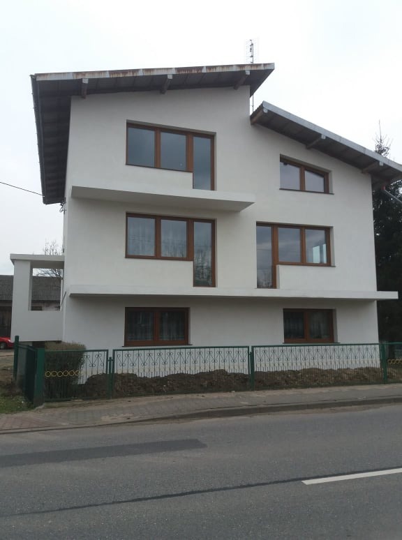 Dwupiętrowy dom z białymi ścianami i brązowymi ramami okiennymi, widziany z poziomu ulicy za zielonym ogrodzeniem.