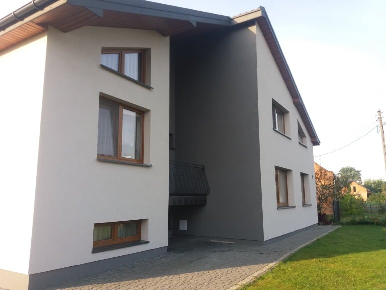 Dwukondygnacyjny dom jednorodzinny z elewacją w kolorze białym i grafitowym, drewniane okna, widok z poziomu chodnika.