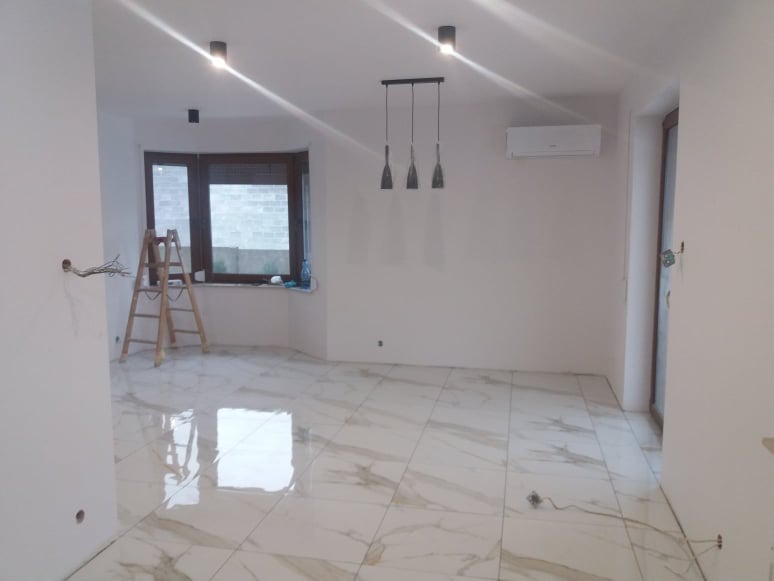 Wnętrze pokoju w trakcie prac wykończeniowych: białe ściany, marmurowe płytki podłogowe z połyskiem, drabina oparta o ścianę pod oknem, wisząca lampa z trzema kloszami, klimatyzator na ścianie...