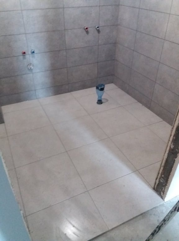 Wykończona kabina prysznicowa z jasnymi płytkami na ścianach i podłodze, widoczne przyłącza wodne i odpływ podłogowy, zabezpieczony plastikową butelką.