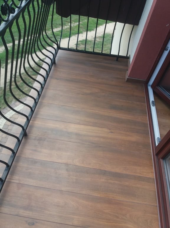 Balkon z podłogą wyłożoną brązowymi panelami imitującymi drewno, czarna metalowa balustrada, widok na zieleń za barierką.