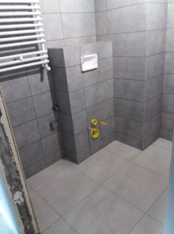 Szara łazienka w trakcie remontu z widocznymi przyłączami hydraulicznymi i grzejnikiem drabinkowym, częściowo wykończona płytkami.