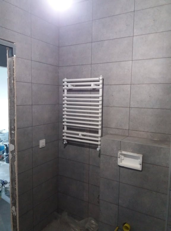 Szare, kwadratowe płytki na ścianie łazienki, biały grzejnik drabinkowy i przygotowane miejsce na toaletę podwieszaną.