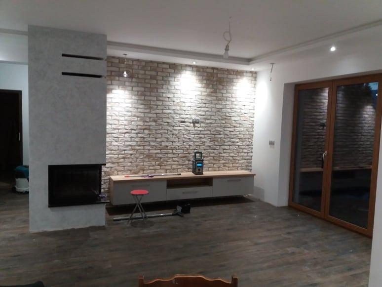 Salon po remoncie: ściana z cegły dekoracyjnej za telewizorem, szary kominek z czarnym wkładem, drewnopodobna podłoga, stolik monterski i niezabezpieczone kable wystające z sufitu.