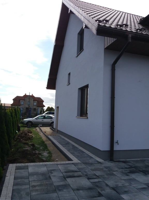 Świeżo otynkowany dom jednorodzinny z grafitową dachówką i rynnami, widok z boku na elewację i chodnik z kostki brukowej, w tle drugi dom i zaparkowany samochód.
