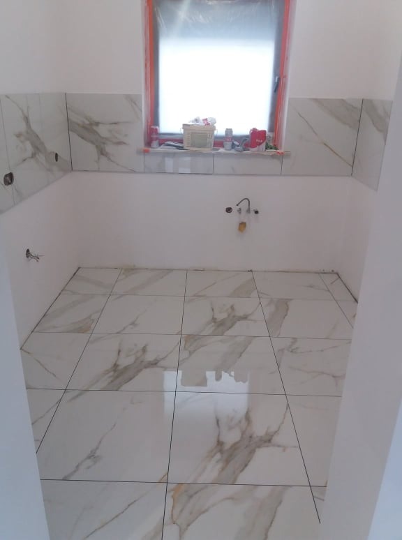 Łazienka w trakcie wykańczania: marmurowe płytki na podłodze i częściowo na ścianach, widoczne otwory na instalacje, okno z folią ochronną.