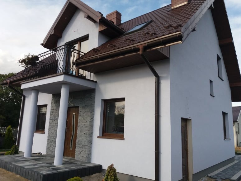 Dwukondygnacyjny dom jednorodzinny z balkonem, wykończony białą elewacją, brązową dachówką i stolarką okienną, z fragmentem elewacji pokrytym dekoracyjnym kamieniem, widok z zewnątrz w pochmurny...