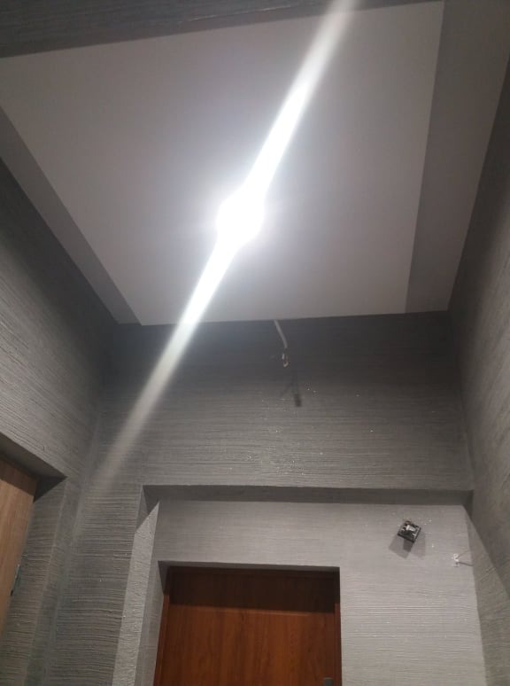 Wykończone wnętrze: Szpachlowane ściany z poziomą teksturą w odcieniach szarości, biały sufit z obniżonym fragmentem i wystającym kablem, widoczny fragment drewnianych drzwi.