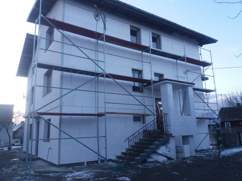Dwupiętrowy dom w trakcie ocieplania, pokryty białymi płytami styropianowymi, otoczony metalowym rusztowaniem, z widocznymi schodami wejściowymi i fragmentem podwórka z resztkami śniegu.