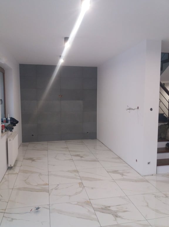 Wnętrze pokoju po remoncie, z białymi ścianami, szarą ścianą z płytek, marmurową podłogą i widocznym fragmentem schodów.