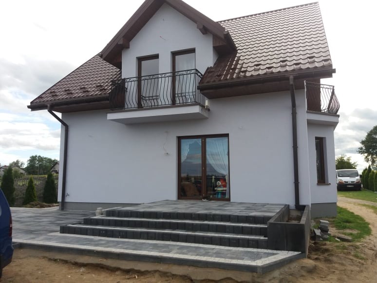 Wykończony dom jednorodzinny z brązowym dachem, balkonem z kutą balustradą i nowym wejściem z kostki brukowej w odcieniach szarości.