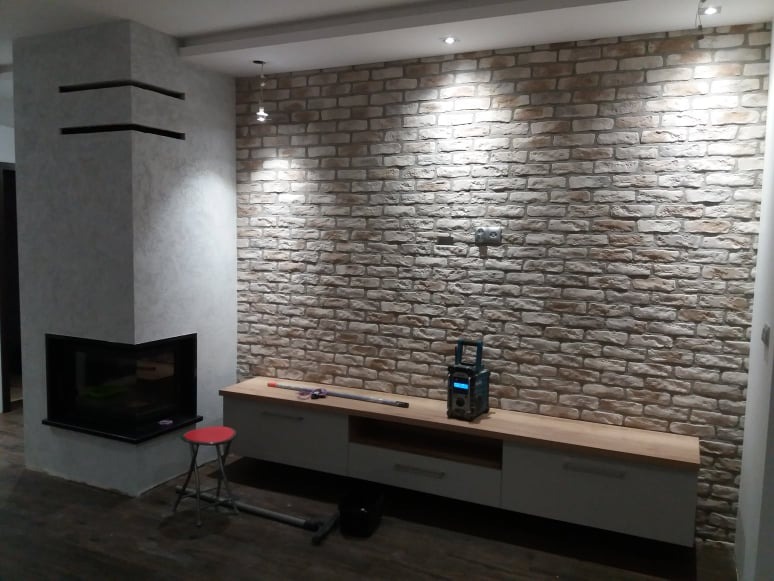 Salon z kominkiem narożnym i ścianą wyłożoną dekoracyjną cegłą, oświetloną punktowymi lampami sufitowymi. Na szafce RTV leżą narzędzia i radio budowlane.