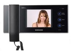 Czarny wideodomofon Samsung z kolorowym wyświetlaczem prezentującym uśmiechniętą kobietę, zamontowany na białej ścianie z widocznym kablem spiralnym.
