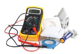 Żółty multimetr cyfrowy z podłączonymi przewodami pomiarowymi, obok białe gniazdko elektryczne, puszka instalacyjna, niebieska taśma izolacyjna i śrubokręt.