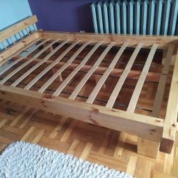 Cezary Buczyński Usługi stolarsko-ciesielskie - Surowa rama łóżka z jasnego drewna z listwami, widoczna na tle parkietu i grzejnika. Elementy łóżka są świeżo obrobione, z widocznymi słojami drewna.