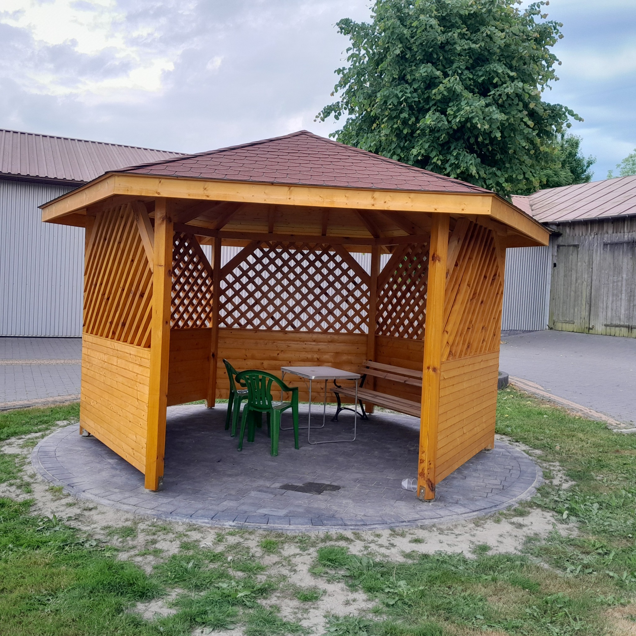 Drewniana altana ogrodowa z ażurowymi ścianami, brązowym dachem i meblami wewnątrz, otoczona kostką brukową i zielenią. Widok z zewnątrz, w tle budynki.