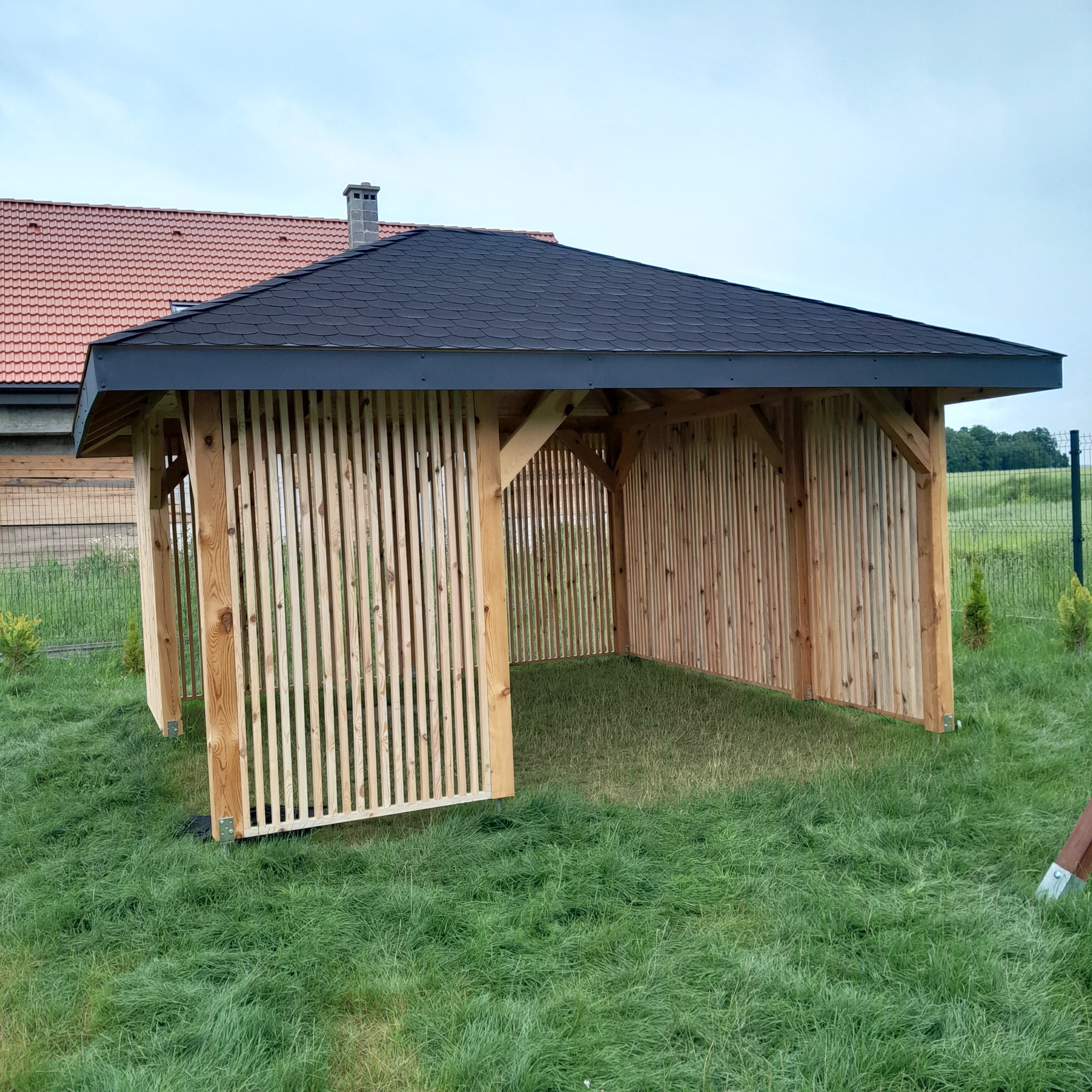 Drewniana altana z czarnym dachem na trawniku, z pionowymi listwami tworzącymi ściany. Widoczne elementy konstrukcyjne i otoczenie zieleni.