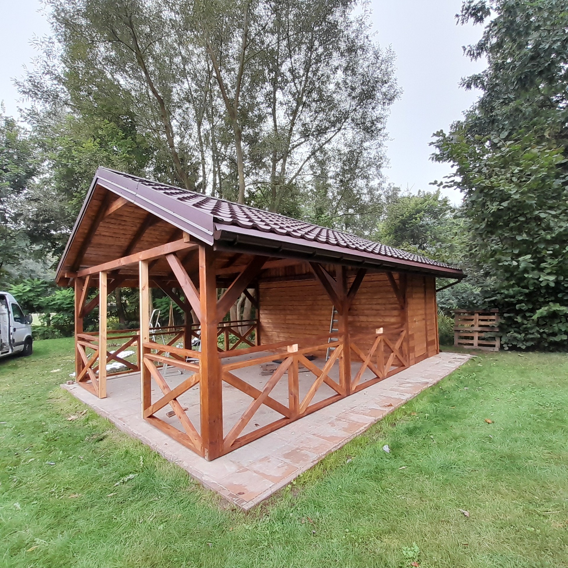 Drewniana altana z brązowym dachem na betonowej podstawie, otoczona zielenią. Widoczna drabina i palety w tle. Konstrukcja z krzyżowymi elementami.