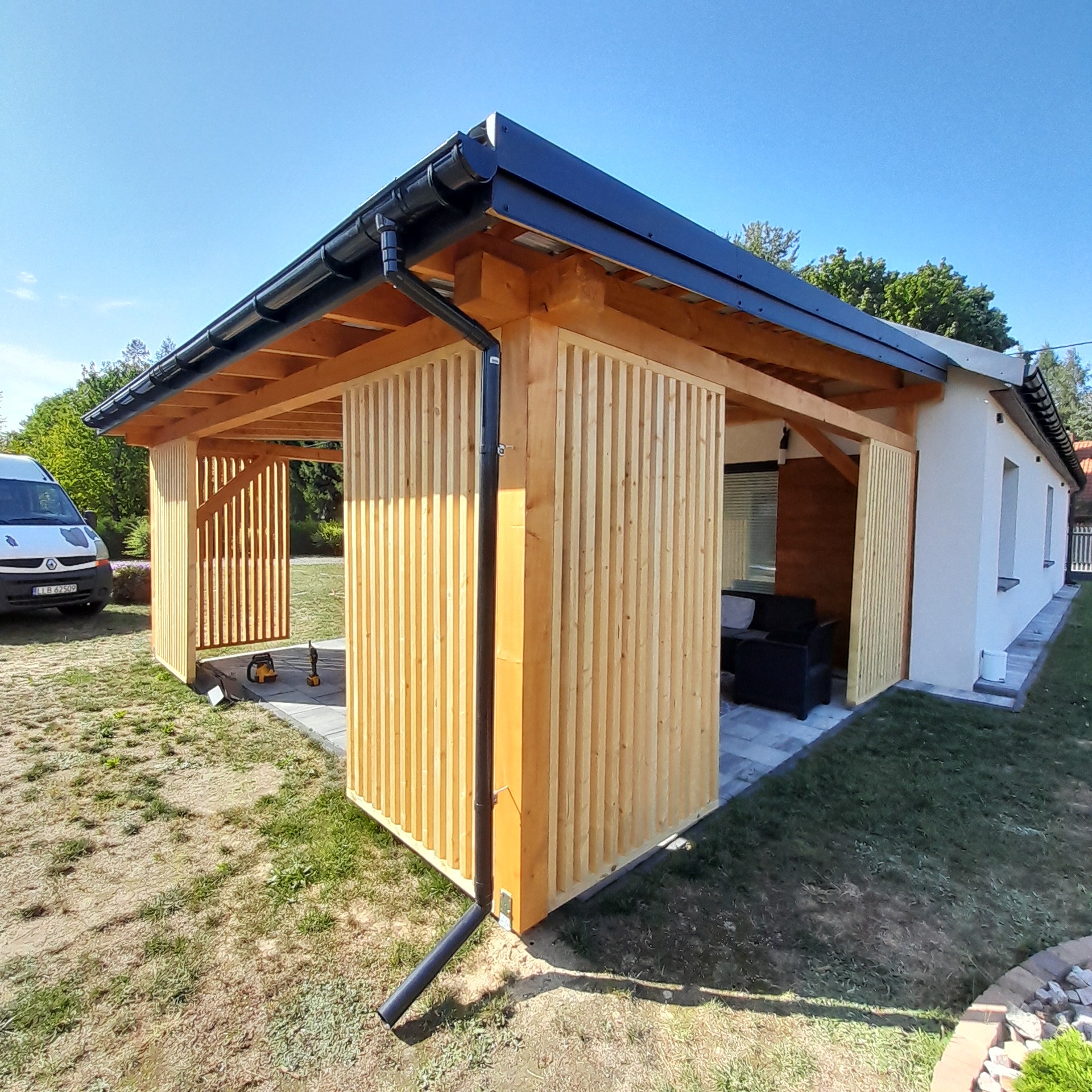 Nowoczesna, drewniana pergola z lamelami, przylegająca do budynku. Widoczne narzędzia i fragment samochodu dostawczego. Słoneczny dzień, trawnik.