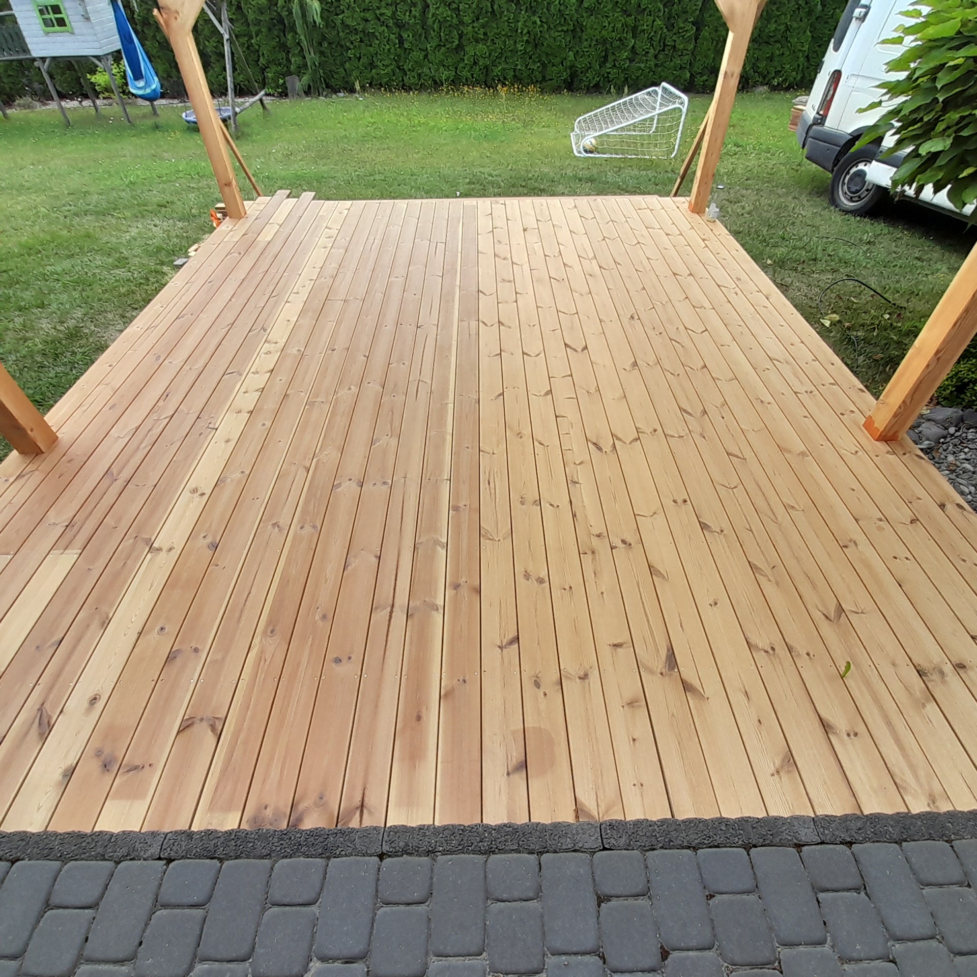 Jasny, drewniany taras w ogrodzie, z widocznymi słupami konstrukcyjnymi i kostką brukową na obrzeżach. W tle trawnik, krzewy i elementy wyposażenia ogrodu.