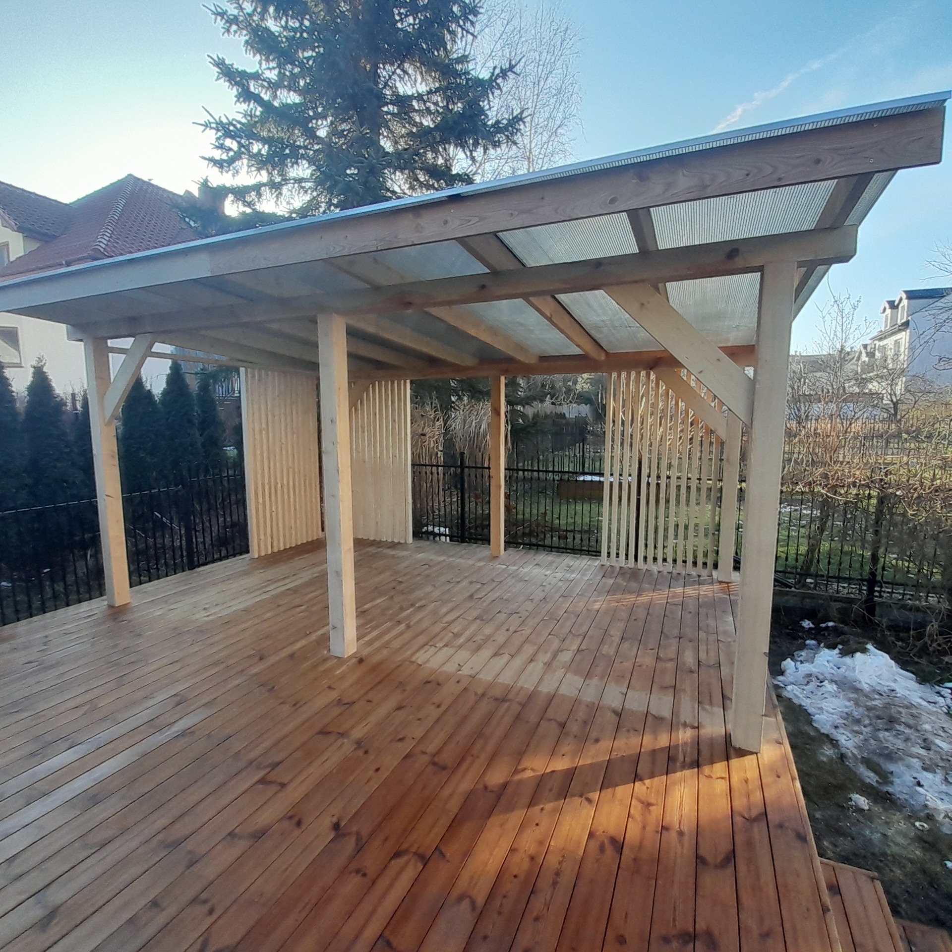 Drewniany taras z zadaszeniem z przezroczystego tworzywa, częściowo osłonięty ażurowymi ściankami. Widoczne drzewa w tle i resztki śniegu.