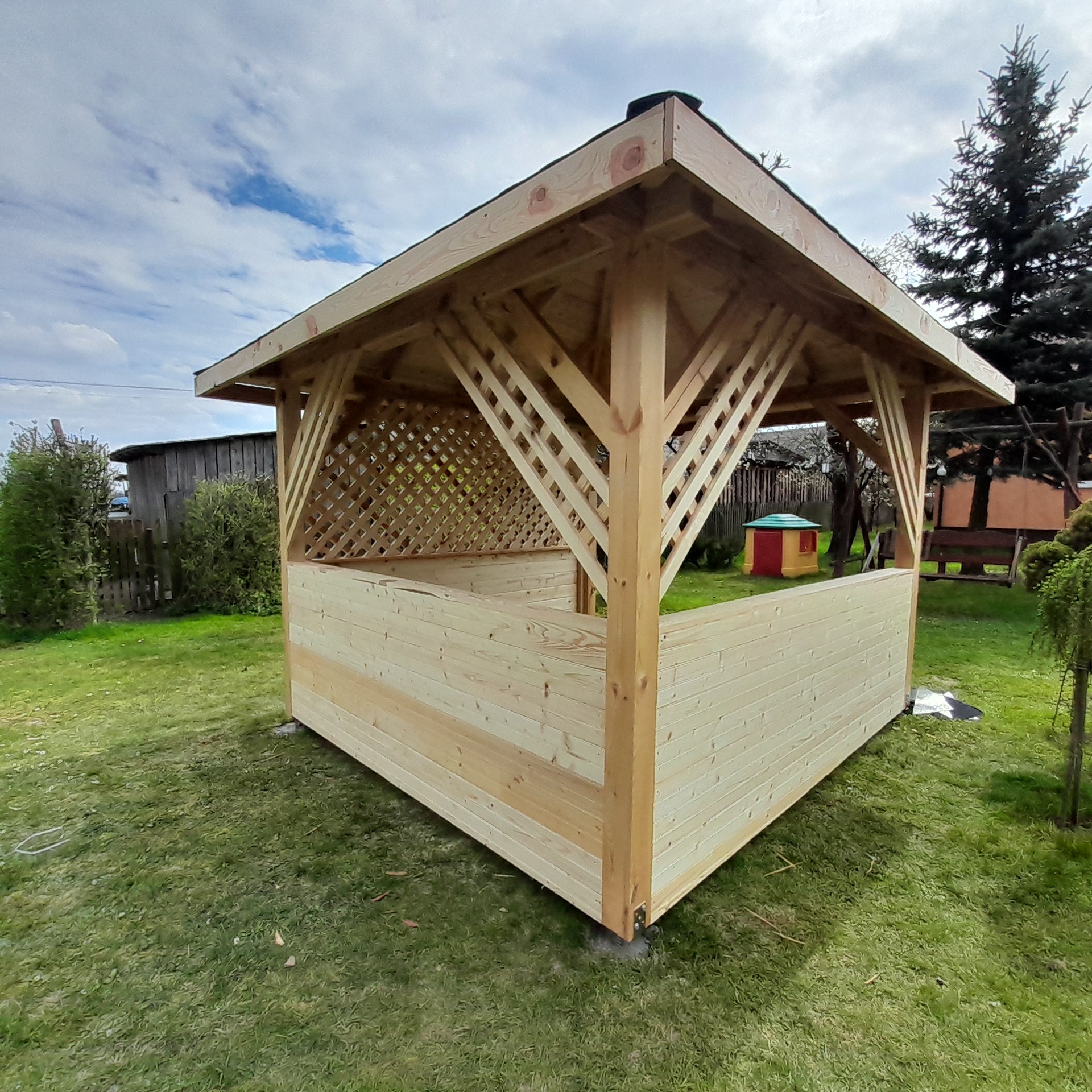 Surowa, drewniana altana ogrodowa z kratownicą, na trawniku, w tle zabudowania gospodarcze i drzewa. Widok z boku, z niskiej perspektywy, podkreślający konstrukcję.