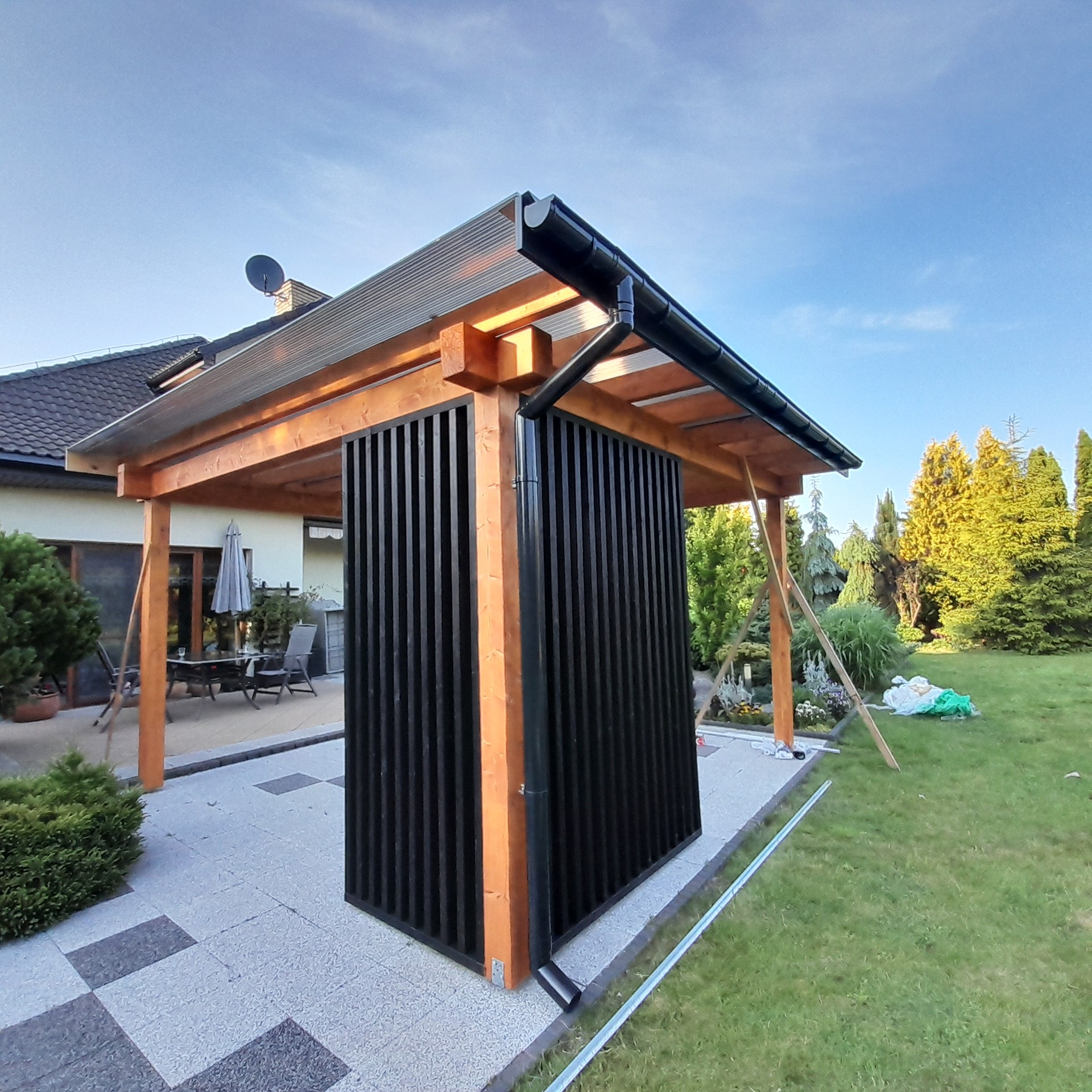 Nowoczesna pergola z drewnianą konstrukcją i czarnymi lamelami, widok z zewnątrz na tle domu i ogrodu. Elementy montażowe widoczne, sugerujące etap końcowy budowy.