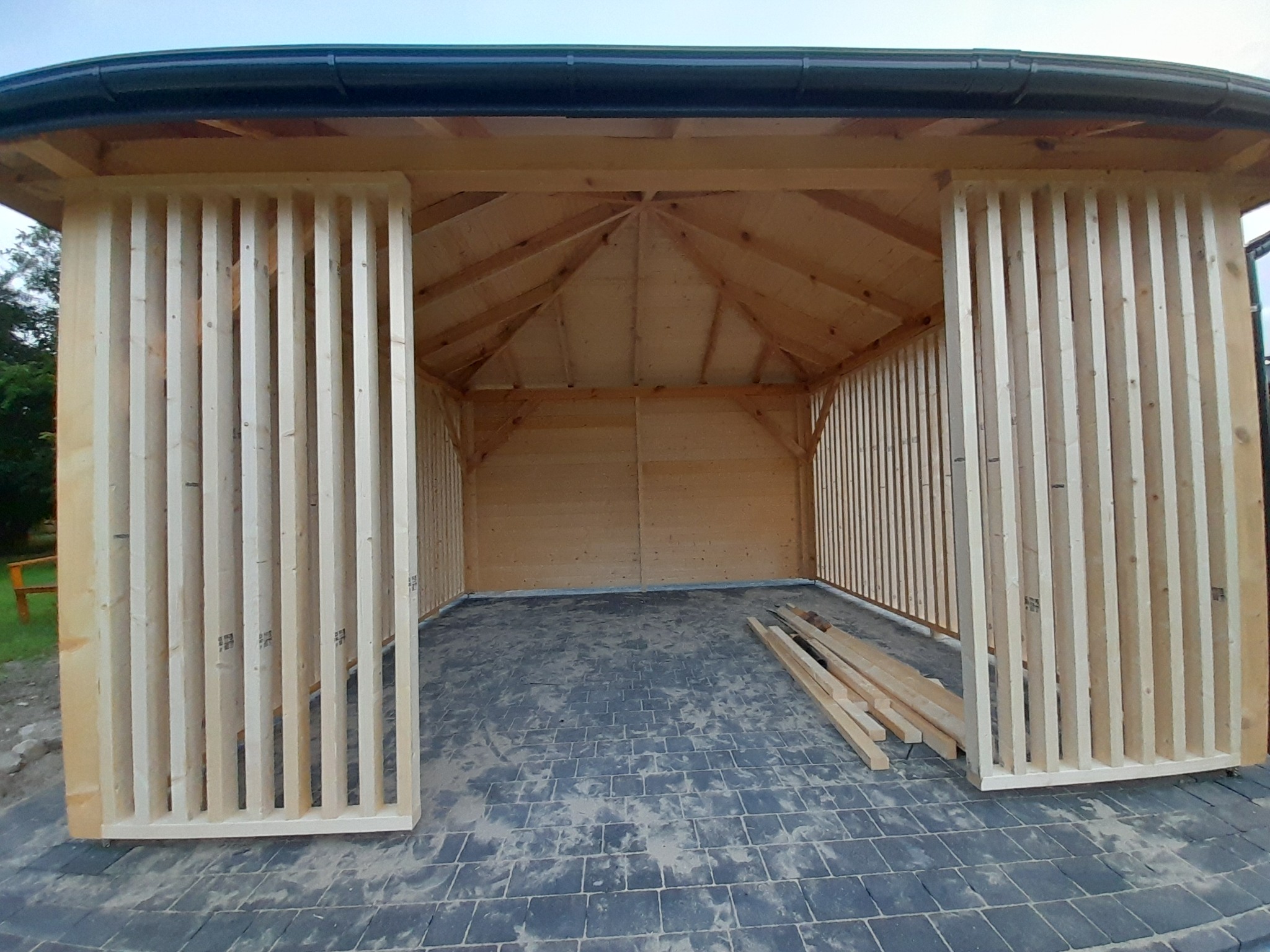 Surowa drewniana konstrukcja altany z lamelami, widok od wewnątrz na szarą kostkę brukową i ułożone deski. Dach z czarną rynną. Etap budowy.