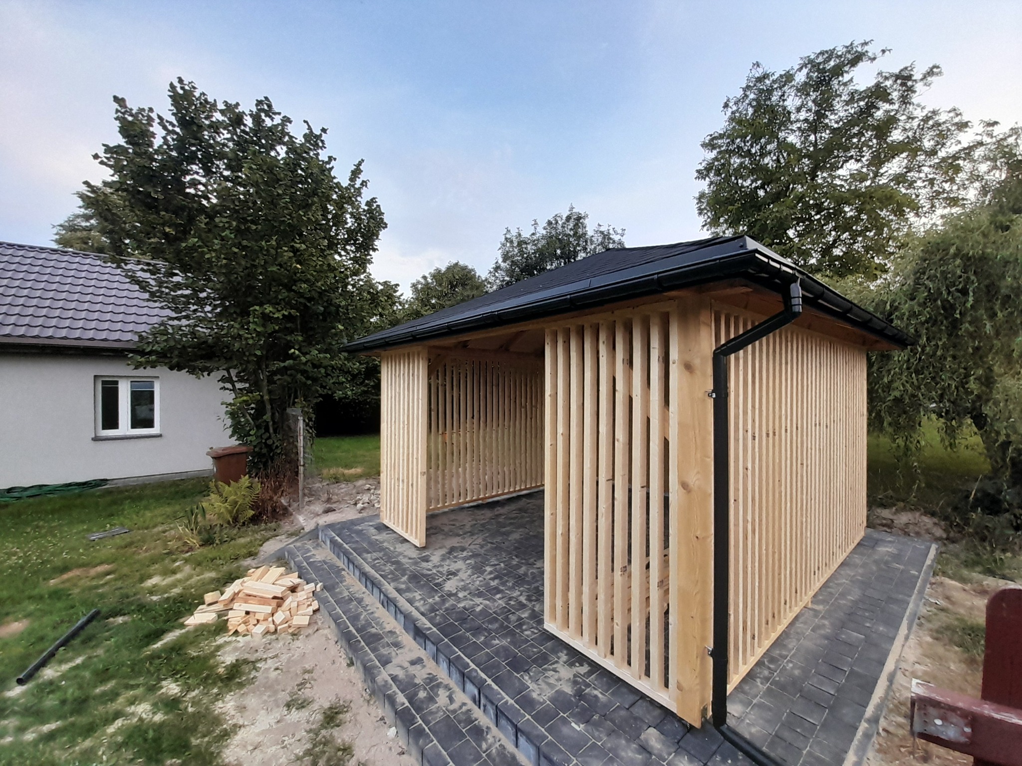Nowoczesna altana z jasnego drewna z pionowymi listwami, czarnym dachem i rynną, na tle domu i zieleni. Ułożona kostka brukowa.