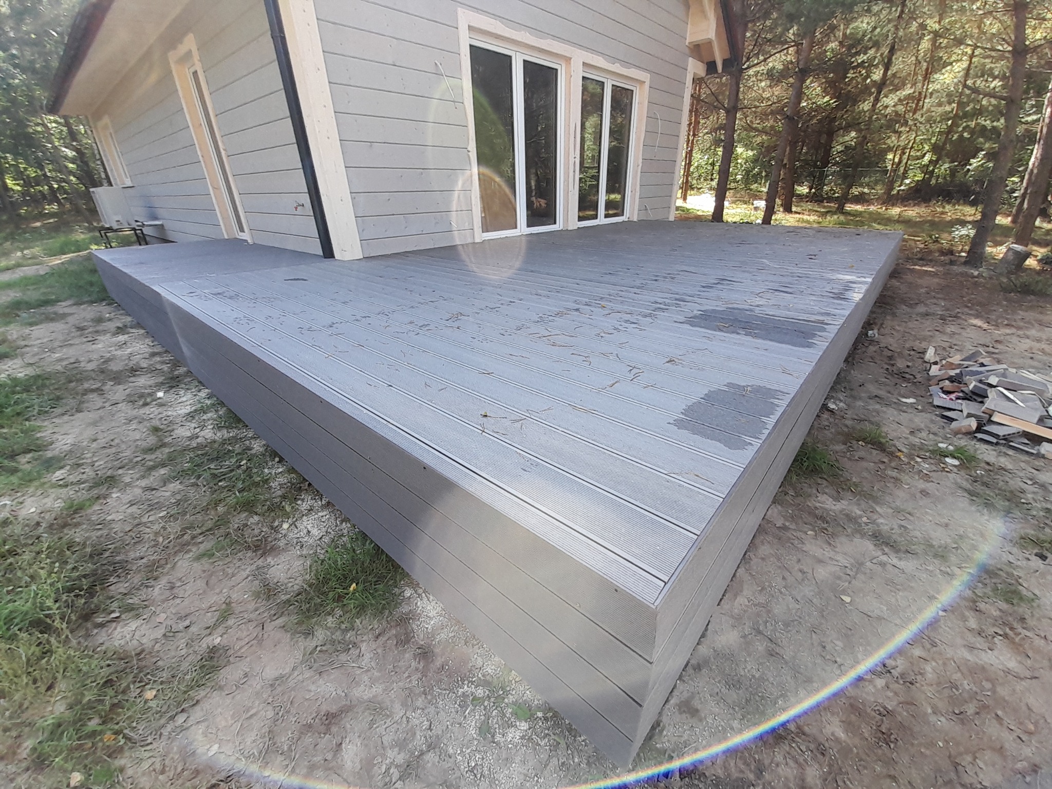 Nowoczesny taras kompozytowy w odcieniach szarości, przylegający do domu z dużymi oknami, otoczony zielenią drzew. Widoczne detale desek i staranne wykończenie.